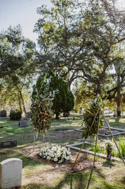 A floral arrangement at a cemetery during daylight, featuring a large bouquet of mixed flowers including yellow, purple, and pink blooms, placed on a tall wooden easel. In front of the arrangement, th