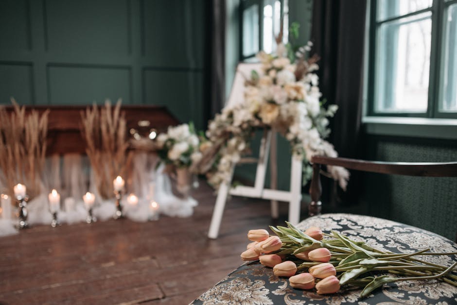 A floral arrangement featuring soft pink tulips with green stems and leaves resting on an ornate, patterned fabric-covered surface in a softly lit room. In the background, a larger display of white an