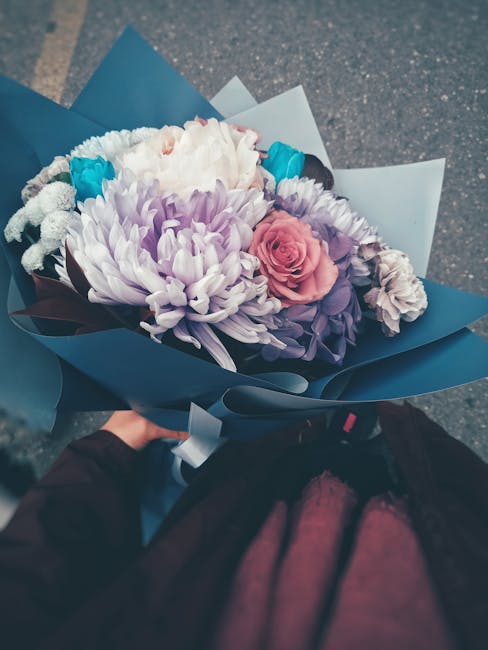 A vibrant bouquet of fresh flowers wrapped in dark blue and light blue paper, featuring large white chrysanthemum blooms, delicate pink roses, smaller purple flowers, and bright blue accent blooms. Th